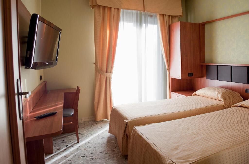 Ein Hotelzimmer mit Schreibtisch, zwei Betten, einem Fernseher und einem Fenster mit Vorhängen.