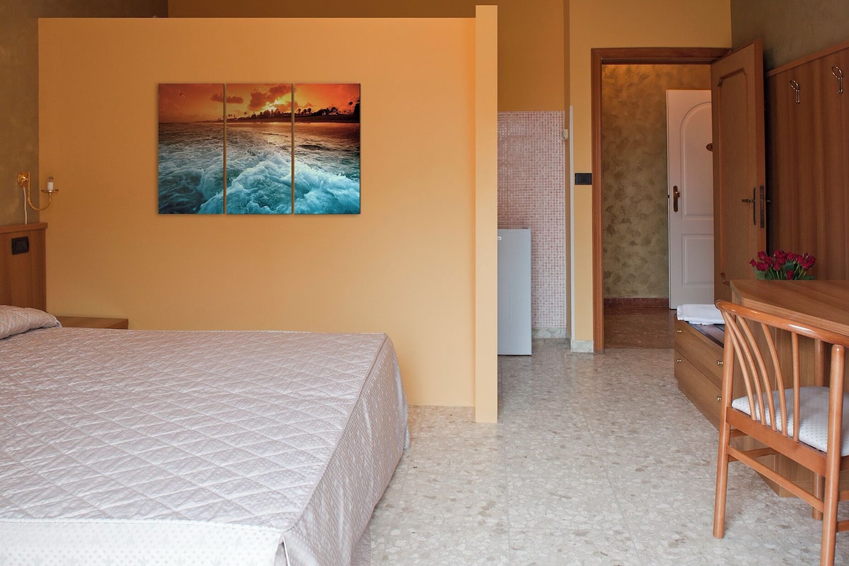 A hotel room with a bed, a desk, a chair, a refrigerator, and a wall-mounted painting.