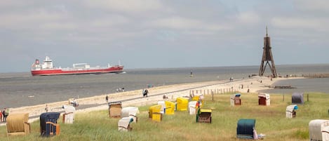 Une plage avec un grand navire en arrière-plan, une structure en bois et plusieurs cabanes de plage numérotées.