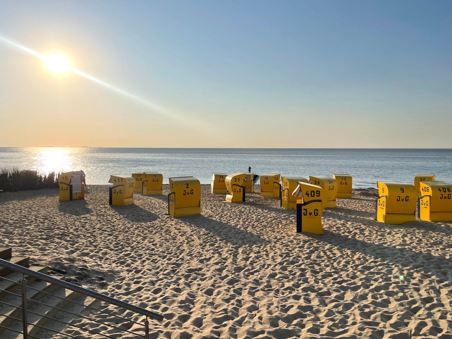 Ein Strand mit gelben Behältern, die mit Nummern beschriftet sind, einer Metalltreppe und dem Meer im Hintergrund.