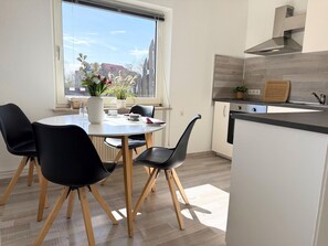A modern kitchen with a dining area, featuring a round table, black chairs, a window with a view, and a built-in stove. - The large Cuxoase - Comfortable vacation apartment with large balcony and close to the beach in (Cuxhaven)
