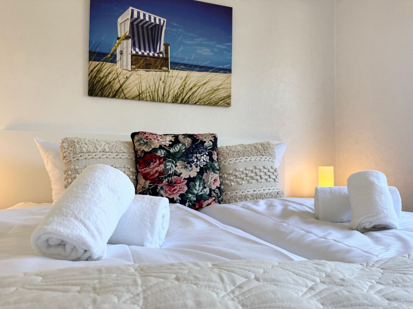 Ein ordentlich bezogenes Bett mit weißen Leinen und Handtüchern, einem Blumenkissen und einem Gemälde mit Strandmotiv an der Wand.