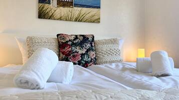 Ein ordentlich bezogenes Bett mit weißen Leinen und Handtüchern, einem Blumenkissen und einem Gemälde mit Strandmotiv an der Wand.