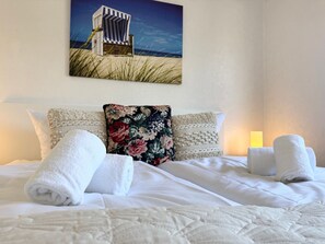 A neatly made bed with white linens and towels, a floral pillow, and a beach-themed painting on the wall. - The large Cuxoase - Comfortable vacation apartment with large balcony and close to the beach in (Cuxhaven)