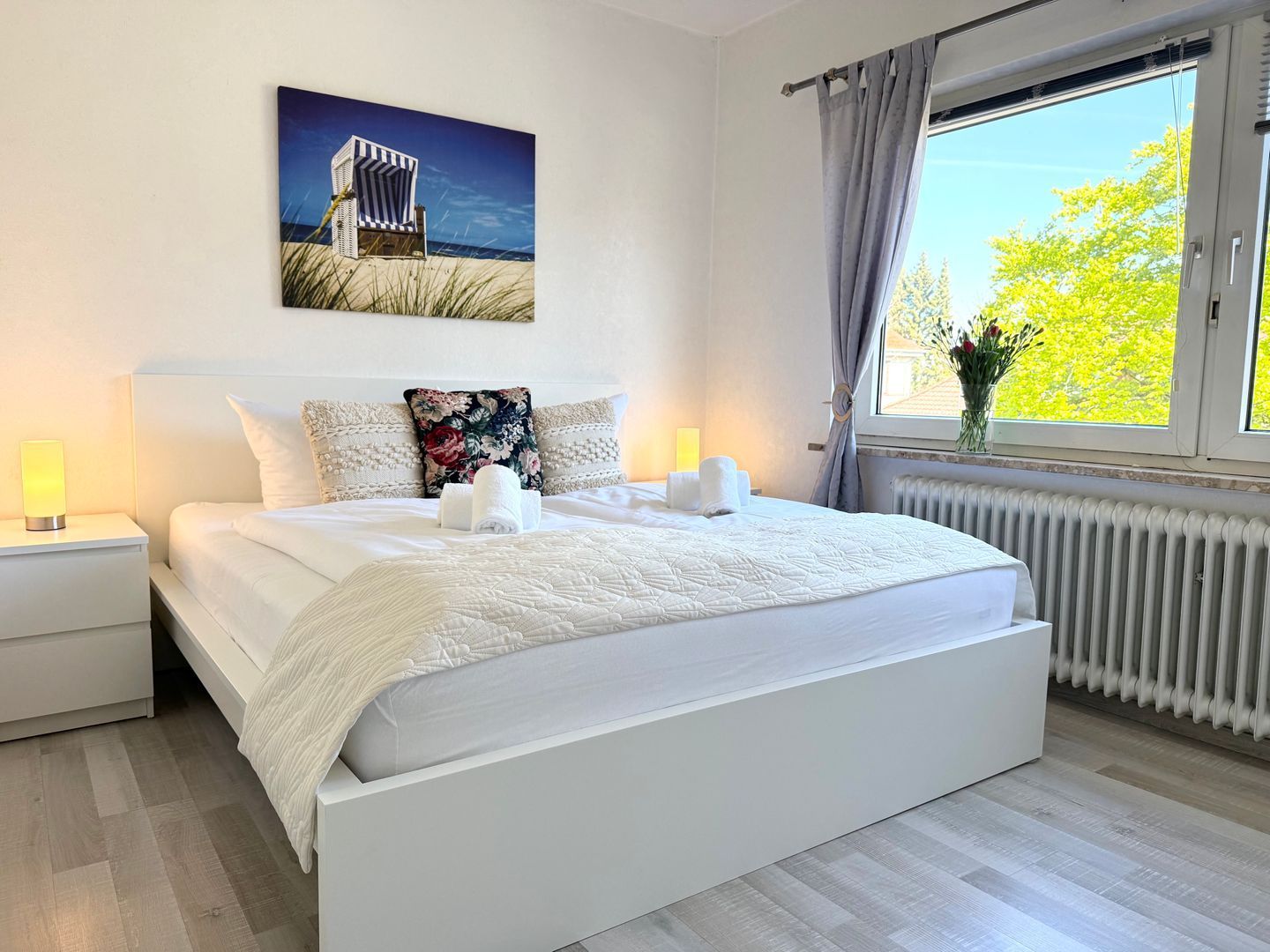 A bedroom with a large bed, two bedside tables, a window with curtains, and a painting on the wall.