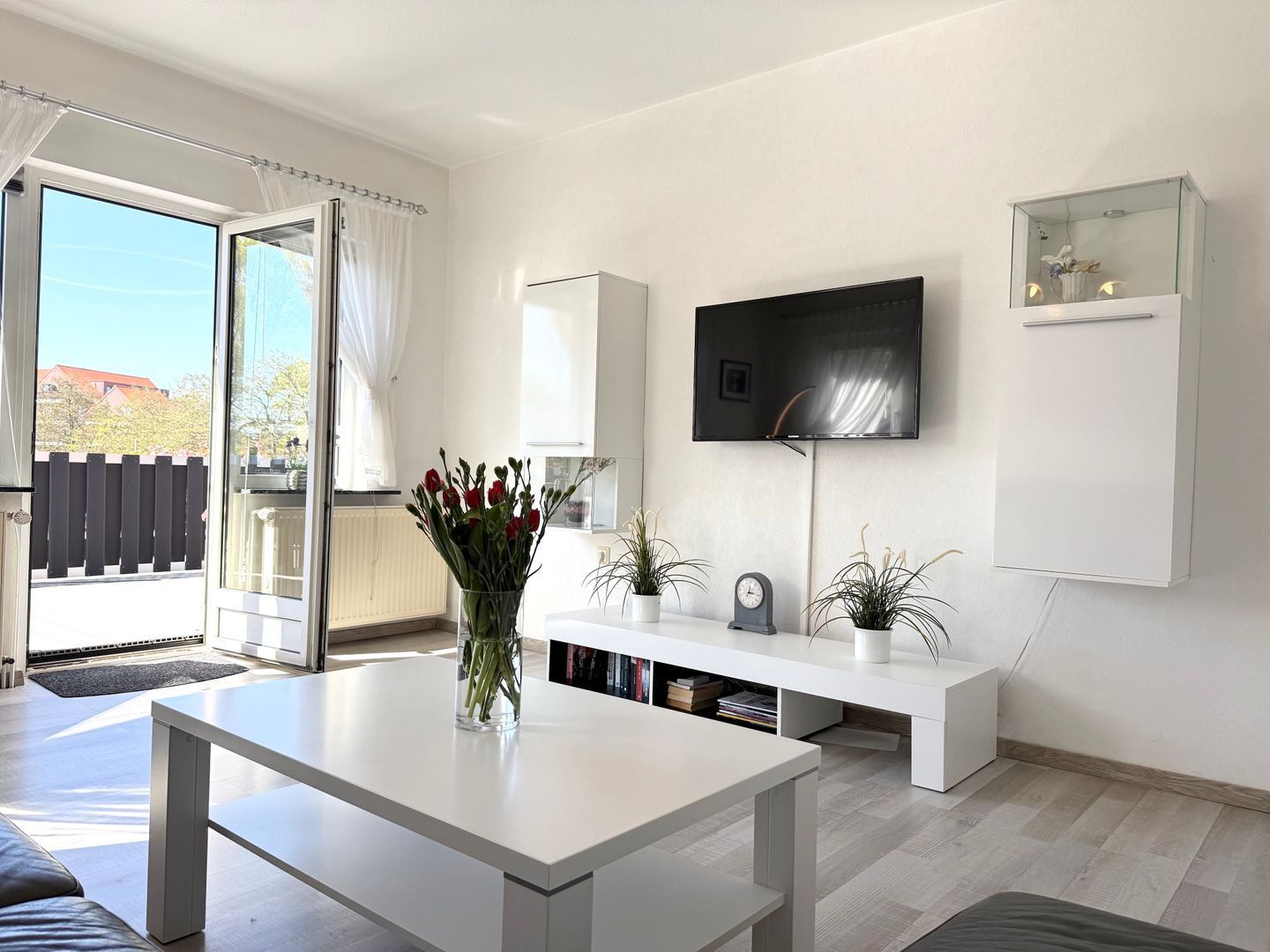 Ein modernes Wohnzimmer mit einem weißen Sofa, einem Couchtisch, einem flachbildfernseher an der Wand, einer Glastür und einer Schiebetür zum Balkon.