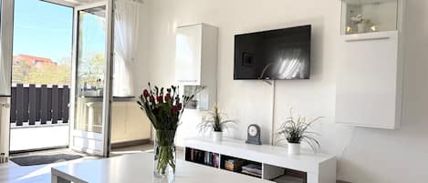Un salon moderne avec un canapé blanc, une table basse, un téléviseur à écran plat fixé au mur, une vitrine et une porte-fenêtre coulissante donnant sur un balcon.