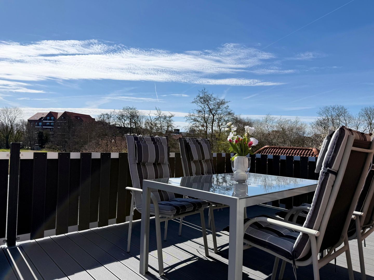 Eine Terrasse im Freien mit einem Glastisch und Stühlen, einer Vase mit Blumen und Blick auf ein Gebäude im Hintergrund.