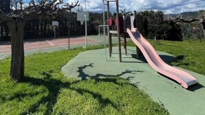 Um parquinho com escorrega e estruturas de escalada, cercado por árvores e uma quadra de basquete.