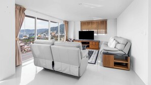 A modern living room with a large window, a flat-screen TV, a sofa, and a wooden cabinet. - Great Ocean View 2Br Near Beach C265 (Patong)