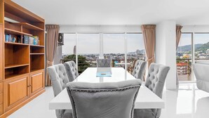 A modern dining area with a white table and grey upholstered chairs, a wooden bookshelf, and large windows offering a city view. - Great Ocean View 2Br Near Beach C265 (Patong)