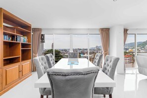Ein moderner Essbereich mit einem weißen Tisch, grau gepolsterten Stühlen, einem Holzregal und großen Fenstern mit Blick auf die Stadt.