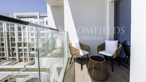 Varanda com cadeiras e mesa redondas de vime, e guarda-corpo de vidro com vista para uma área residencial.