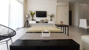 A modern living room with a flat-screen TV mounted on a white wall, a black coffee table, a white sofa, and a dining area with a white chair. - Borneo Holiday Suite @ Riverine Diamond Kuching (Kuching)