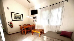 Una sala de estar con un sofá, una mesa de comedor con sillas, un televisor de pantalla plana montado en la pared y un extintor en la pared.