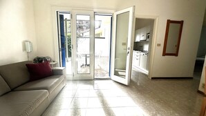 Una sala de estar con un sofá, un ventanal de vidrio que da a un balcón, y una cocina visible a través del ventanal.