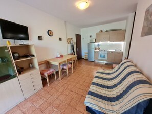 A compact living space with a bed, a desk, a television, and a kitchenette. - Comfortable flat with private garden near the sea (Bibione Pineda)