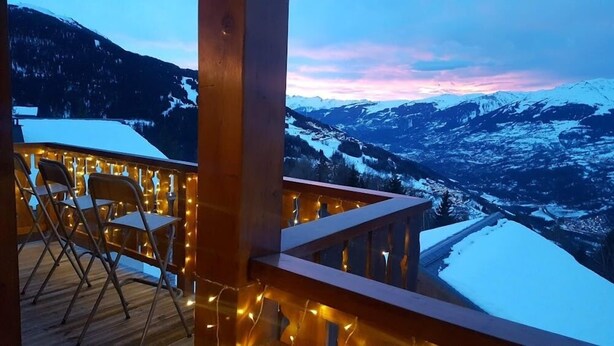 Een houten terras met stoelen en lichtslingers, met uitzicht op een besneeuwd berglandschap.