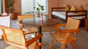 A cozy room with a round wooden table surrounded by four chairs, a hanging light fixture, a bed with a white mattress and yellow pillows, and a potted plant near the window. - Casa Mezcaleria (Boracay Island)