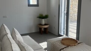 A modern, minimalist living room with a grey sofa, a small round table with a plant, and a large sliding glass door leading to an outdoor area. - Villa Mae by ΜΡS (Agkeria)