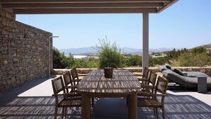 A covered outdoor patio with a stone wall, a wooden table with chairs, and a view of the landscape. - Villa Mae by ΜΡS (Agkeria)