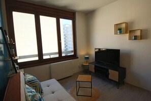 Ein Wohnzimmer mit einer Couch, einem Fernseher, einem kleinen Tisch mit Lampe und einem großen Fenster mit Blick auf Gebäude.