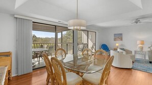 Un salon moderne avec une table à manger en verre entourée de chaises en osier, un canapé blanc et un fauteuil bleu. On aperçoit un ventilateur de plafond et une lampe à l’arrière-plan.