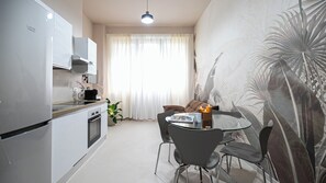 Una cocina compacta con electrodomésticos modernos, un comedor con mesa y sillas de vidrio, y un amplio ventanal con cortinas de gasa.