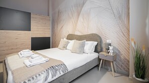 Une chambre d’hôtel moderne avec un grand lit, une table de chevet avec une lampe et un ours en peluche, et un mur orné d’un motif de feuilles.