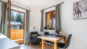 A room with a wooden table and chairs, a window with curtains, a chair with a cushion, and a view of a wooden deck outside. - Apartment 'Fewo Am Steinbach - Stoaboch' with Mountain View, Balcony and Wi-Fi (Reit im Winkl)