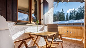 A wooden deck with a table and chairs, a window with curtains, and a view of a snow-covered building and trees. - Apartment 'Fewo Am Steinbach - Stoaboch' with Mountain View, Balcony and Wi-Fi (Reit im Winkl)