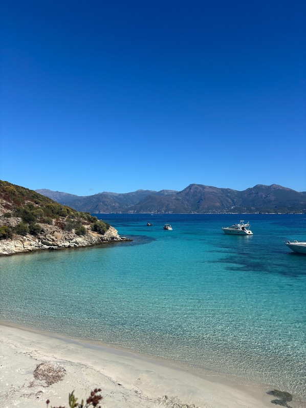 A coastal landscape with clear turquoise waters, boats, and distant mountains.