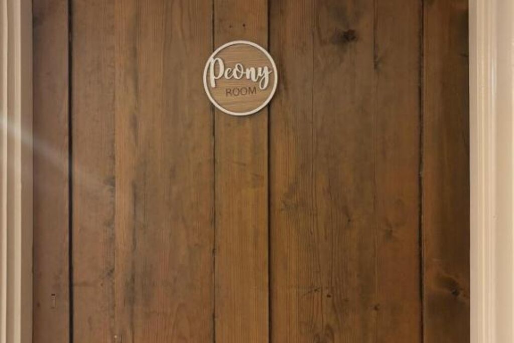 Een houten wand met een rond bord waarop 'Peony Room' staat.