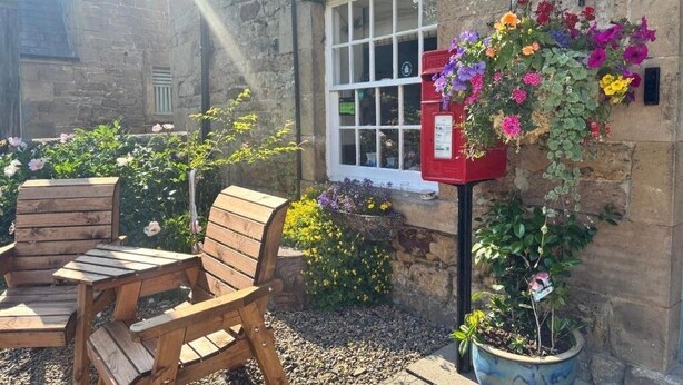 Un bâtiment en pierre avec une fenêtre, une boîte aux lettres rouge, des chaises en bois et un panier à fleurs suspendu.