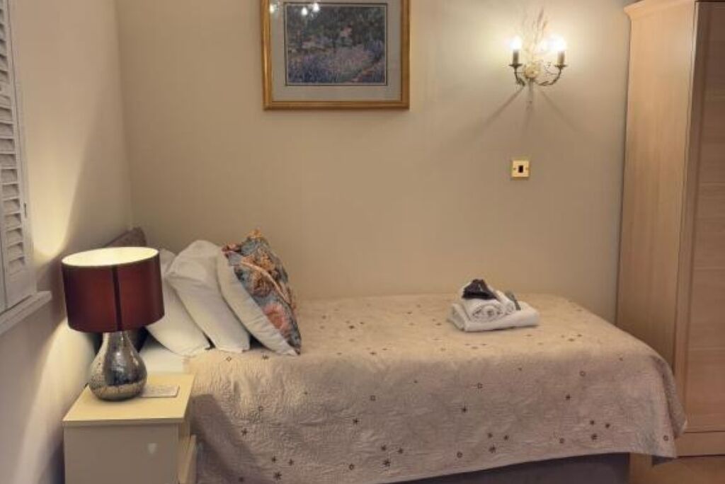 A bedroom with a bed, a nightstand with a lamp, a wall-mounted light fixture, and a framed picture on the wall.