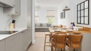 Une cuisine moderne avec des meubles blancs, un coin repas avec une table et des chaises en bois, et un salon avec un canapé et une fenêtre.
