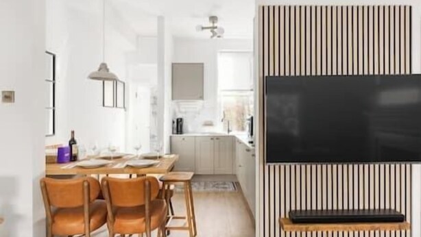 Une cuisine moderne avec des meubles blancs, un coin repas avec des chaises en bois et un grand téléviseur à écran plat fixé sur un panneau en bois.