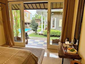 Une chambre d’hôtel avec un lit, une table à manger agrémentée d’un panier de fruits, et une vue sur une piscine et un jardin.