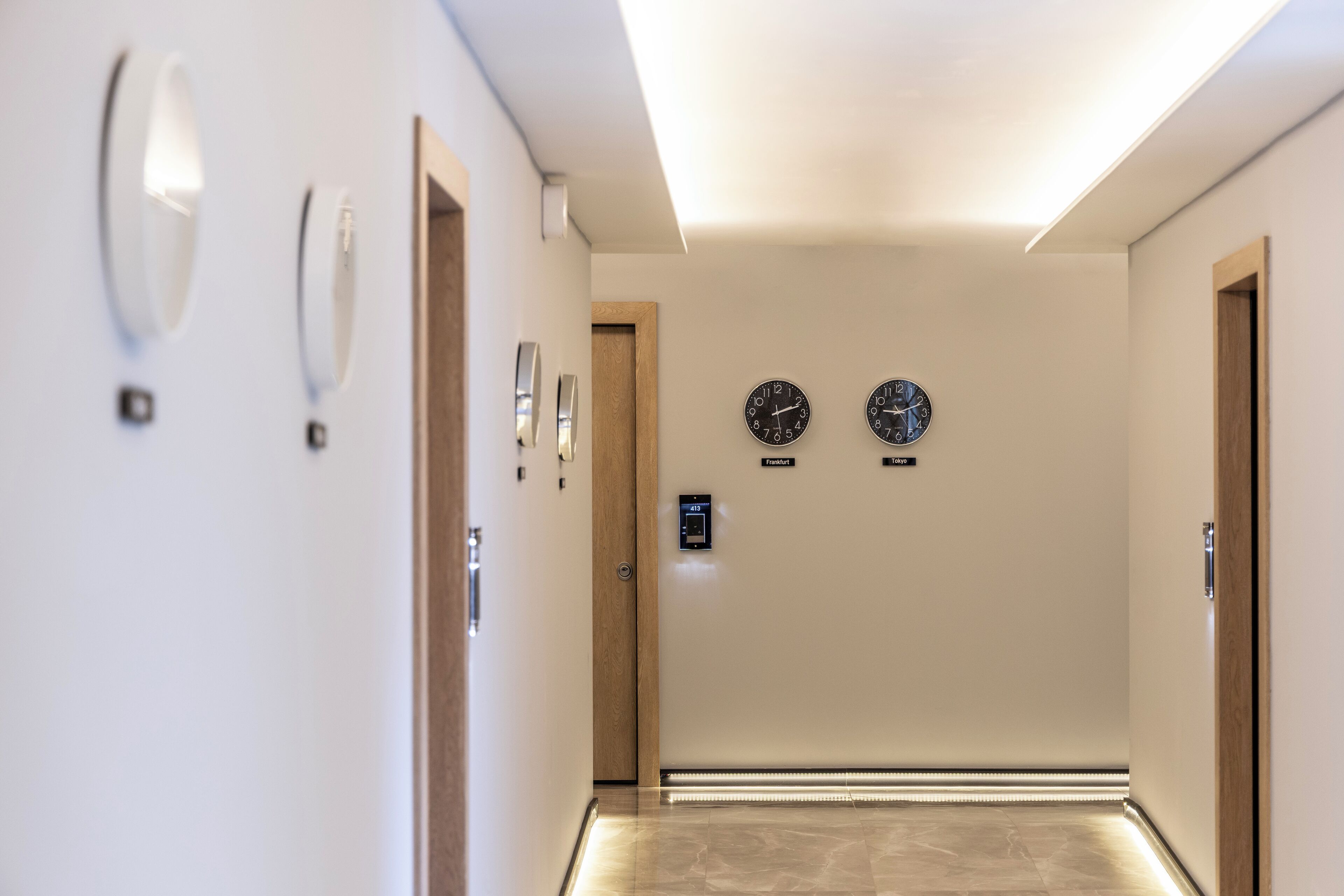 Un couloir d’hôtel moderne avec un sol de couleur claire, une porte en bois au fond et deux horloges accrochées au mur.