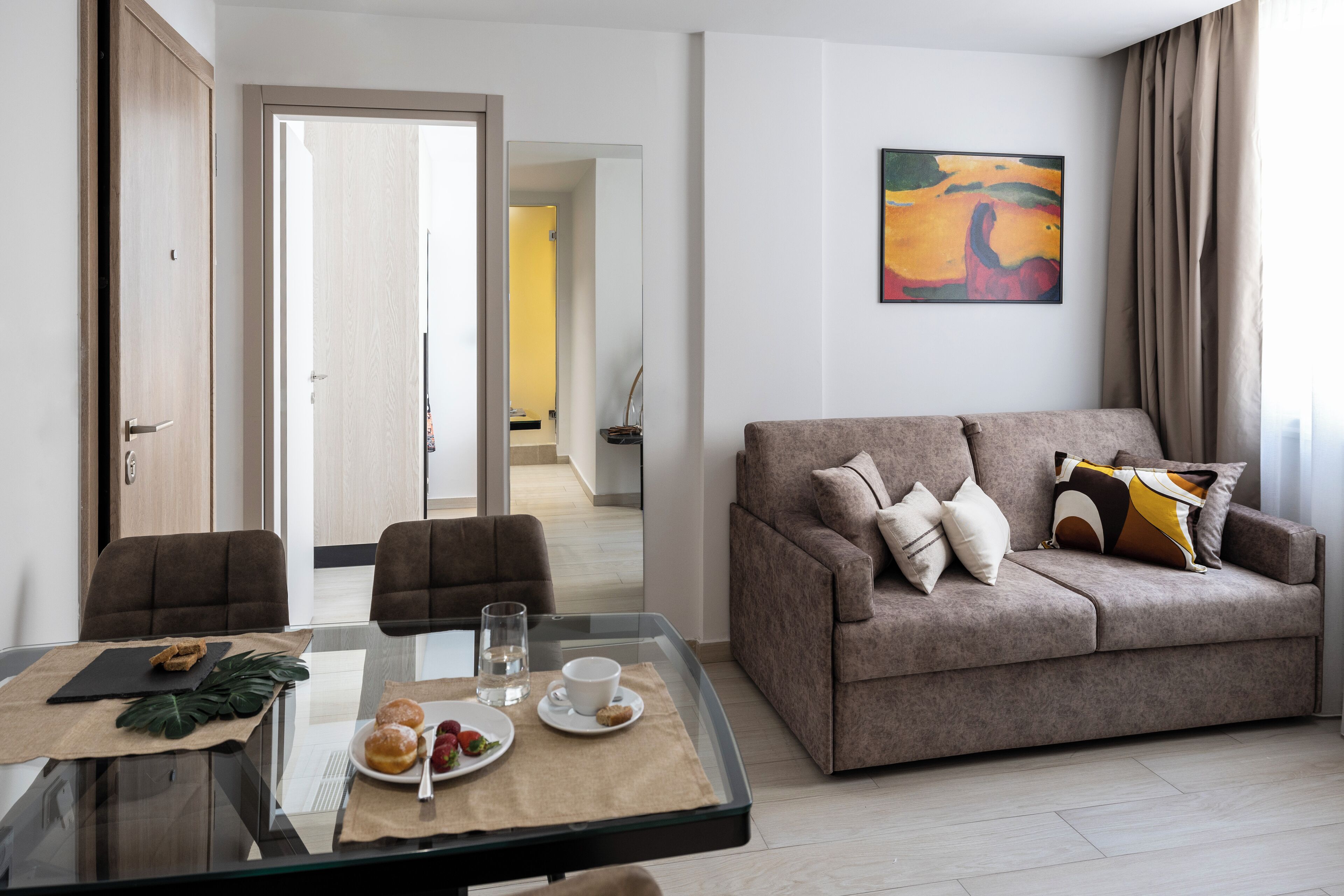 A modern living room with a glass dining table, a sofa, and a painting on the wall.