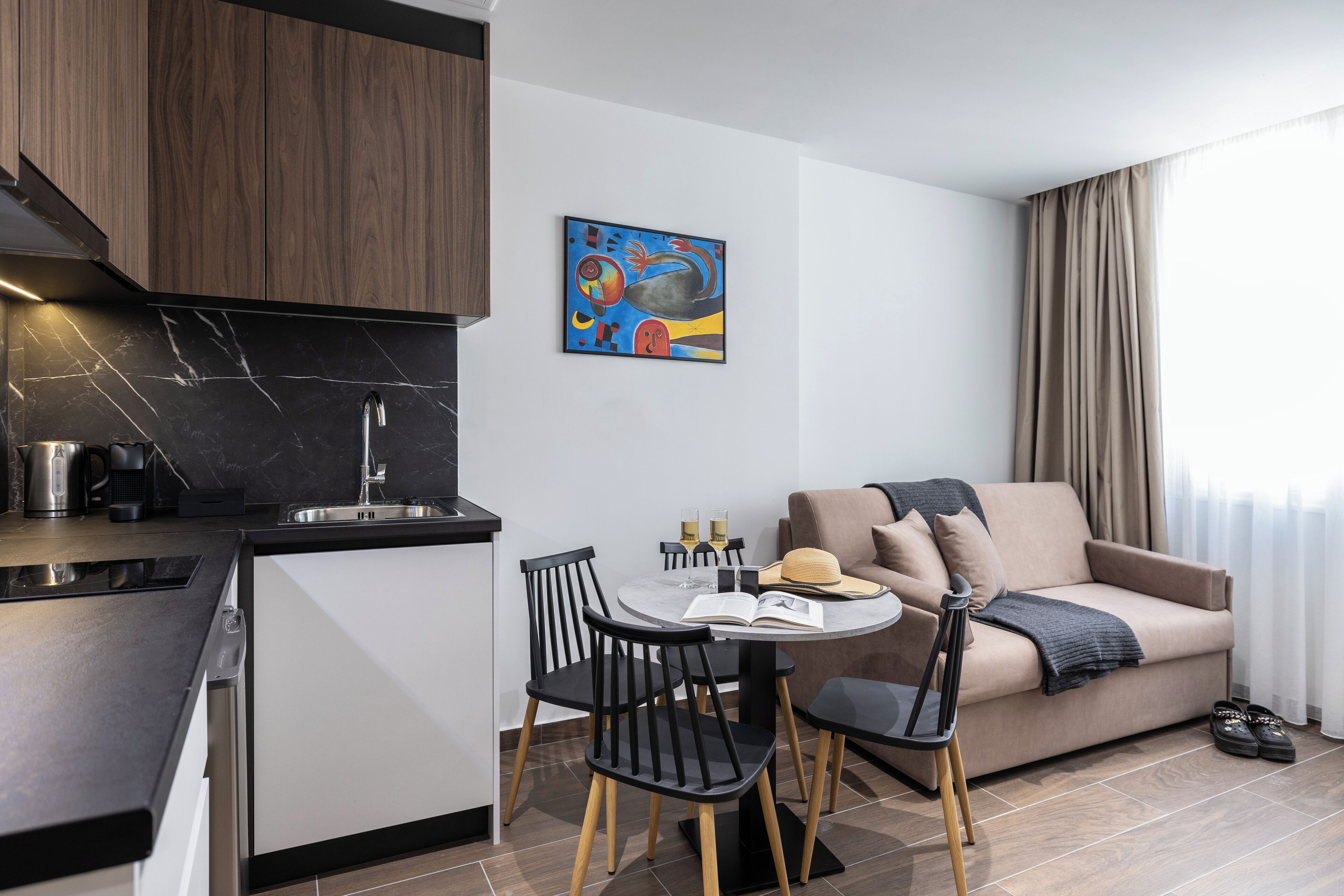A modern kitchen with a black countertop, a built-in stove, and a sink. There is a dining area with a table, chairs, and a sofa. A painting is on the wall, and a window with curtains is visible.