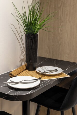 A dining table with a black marble top, a black chair, a woven placemat, a white plate, and a black vase with green plants.