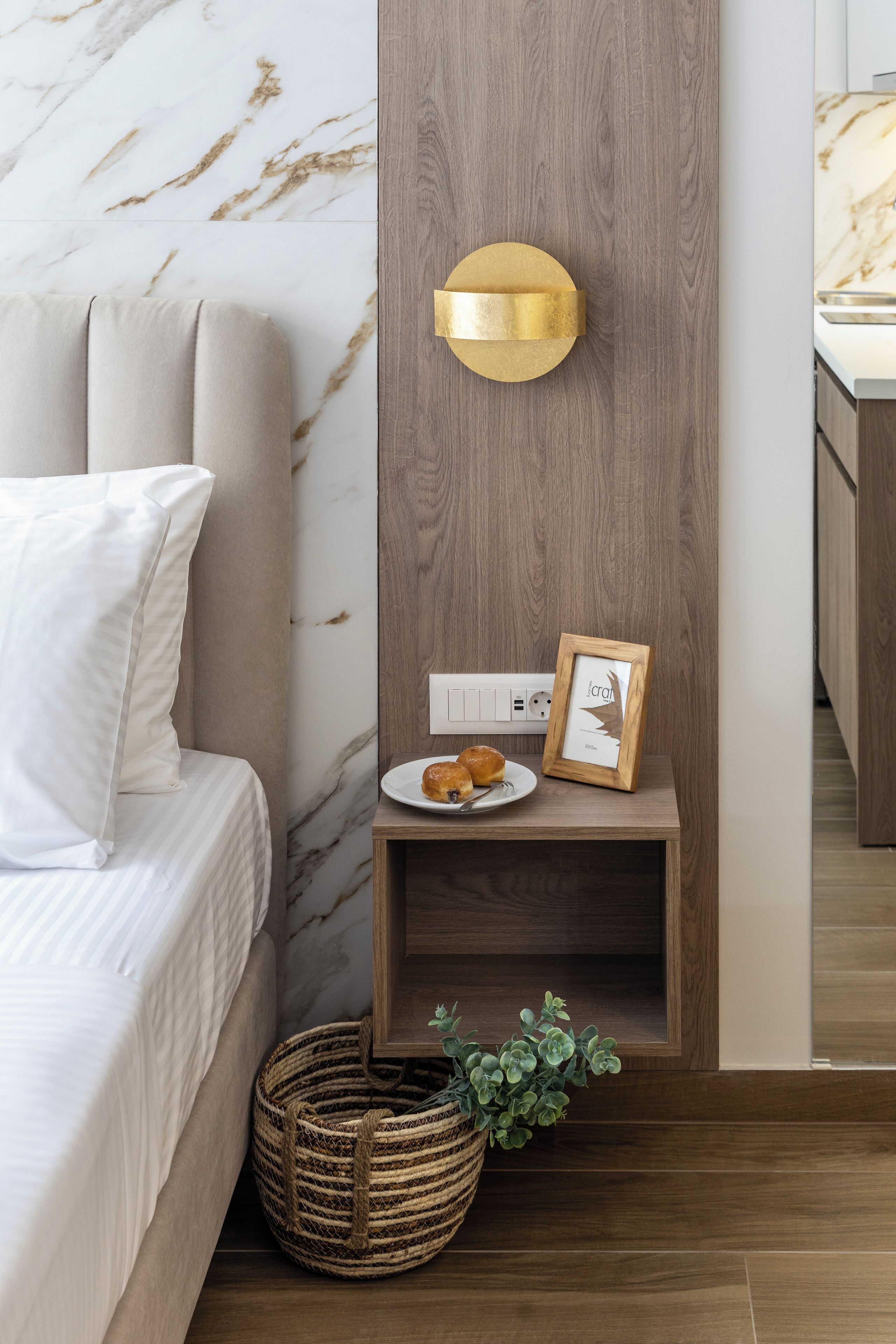 Une chambre moderne avec un mur en marbre, une table de chevet en bois ornée d’un cadre photo, une lampe de chevet et un panier en osier.