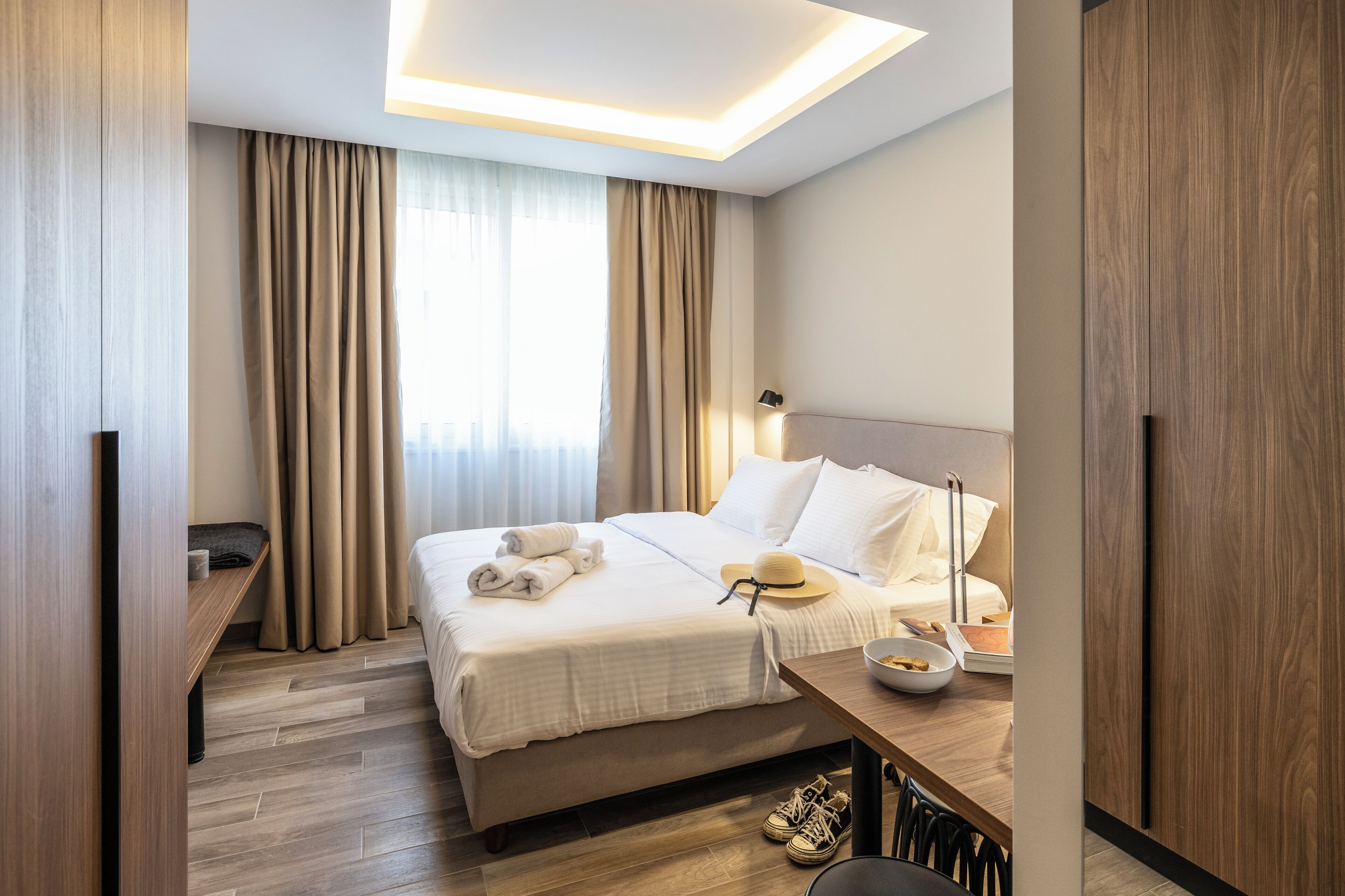 Een moderne hotelkamer met een opgemaakt bed, een bureau met een laptop, en uitzicht op de stad door grote ramen.