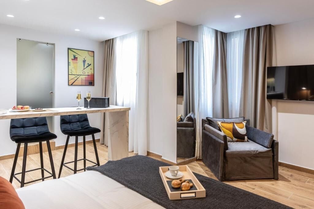 A modern hotel room with a bed, a desk with two bar stools, a sofa, a TV, and a window with curtains.