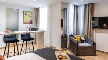 A modern hotel room with a bed, a desk with two bar stools, a sofa, a TV, and a window with curtains.