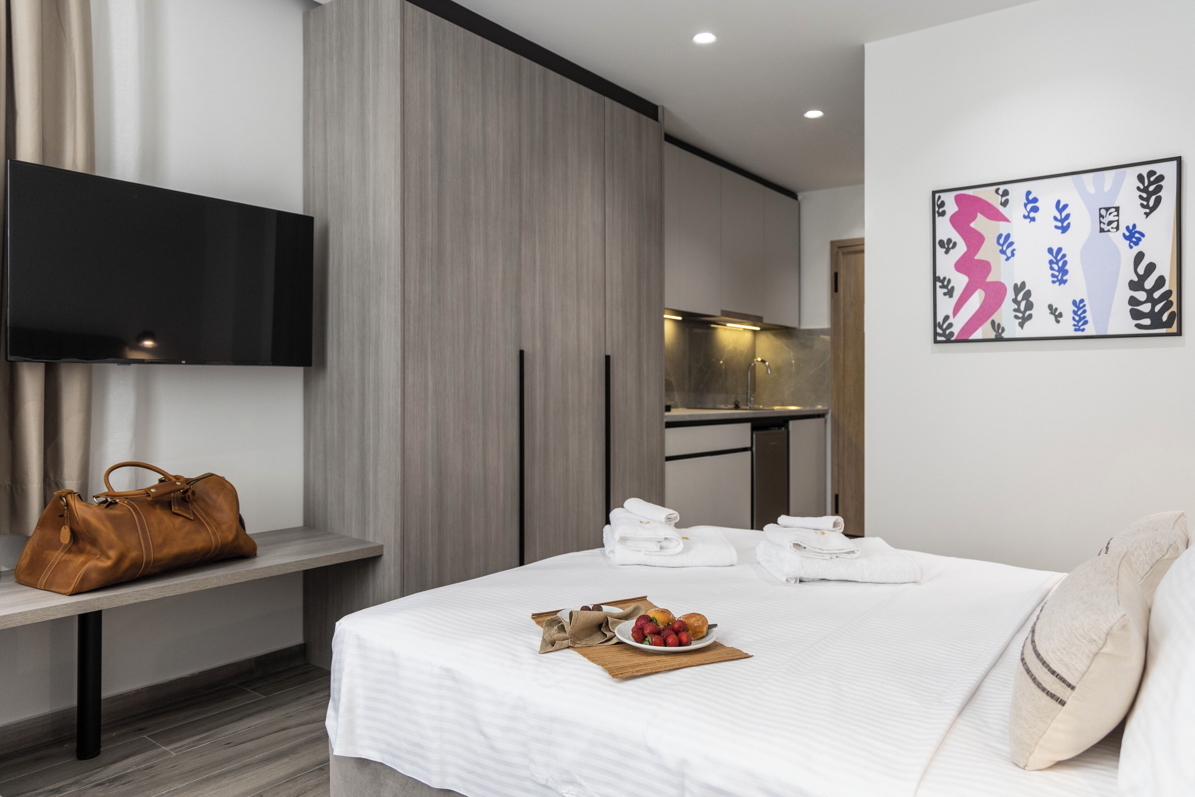 A modern hotel room with a bed, a TV mounted on the wall, a desk with a chair, and a brown leather bag on a bench.