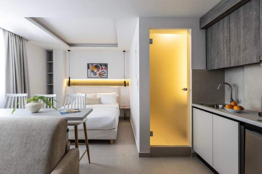 A modern hotel room with a bed, bedside table, and a small kitchenette.