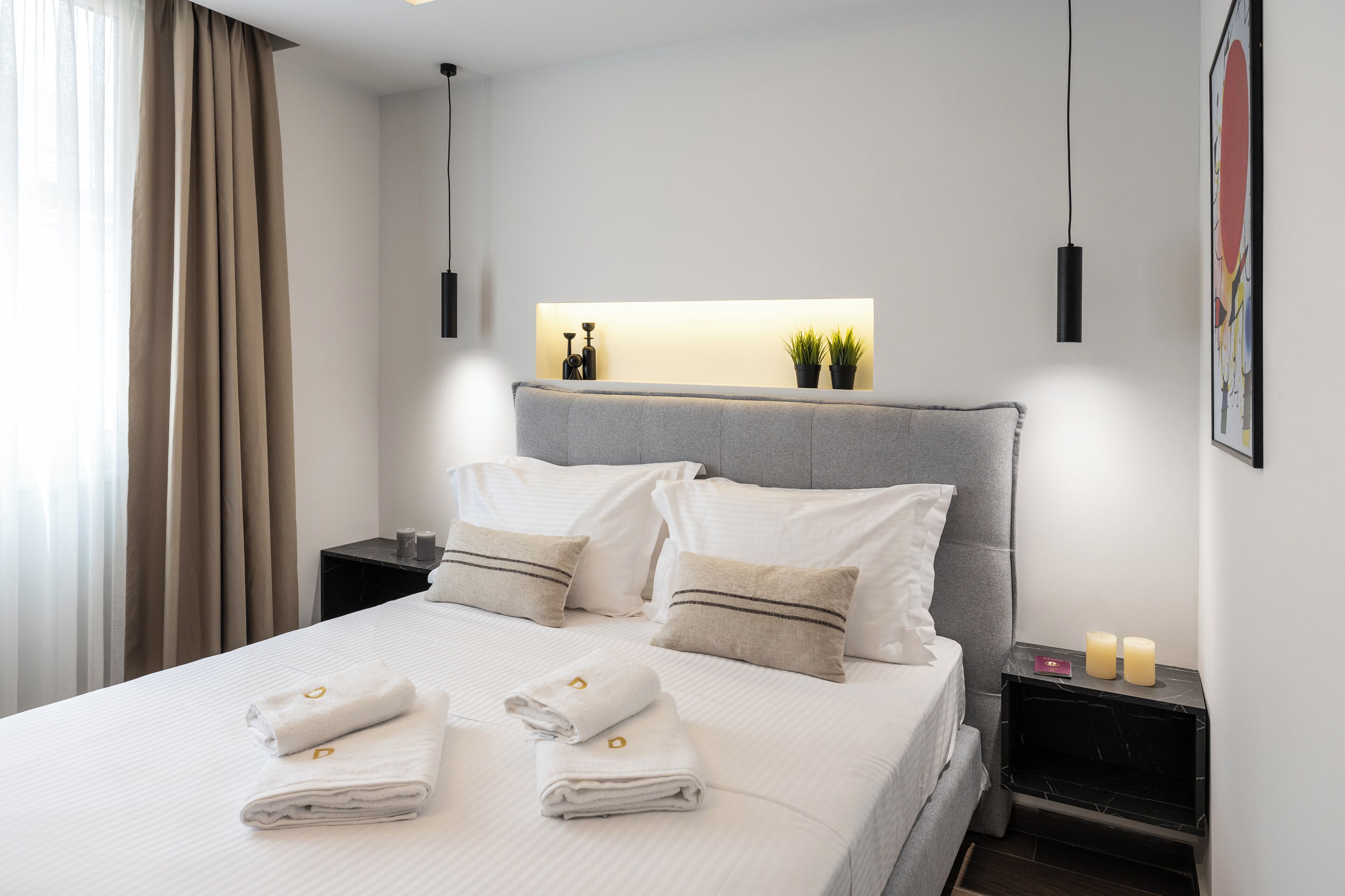 A neatly made bed with white linens and towels, a bedside table with a lamp, and a wall-mounted shelf with decorative items.