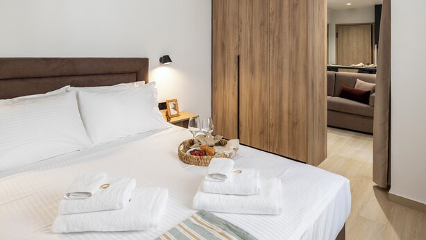 A hotel room with a large bed, a wooden wardrobe, a small table with a lamp, and a basket of fruit and pastries. - DOMIOS Suites Athens (Athens)